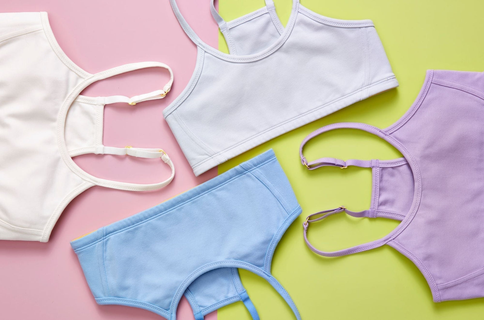 Four Yellowberry Bras styled on a two-color background. The cotton bras are white, light blue, pale blue, and lighter purple. The background is green and pink.