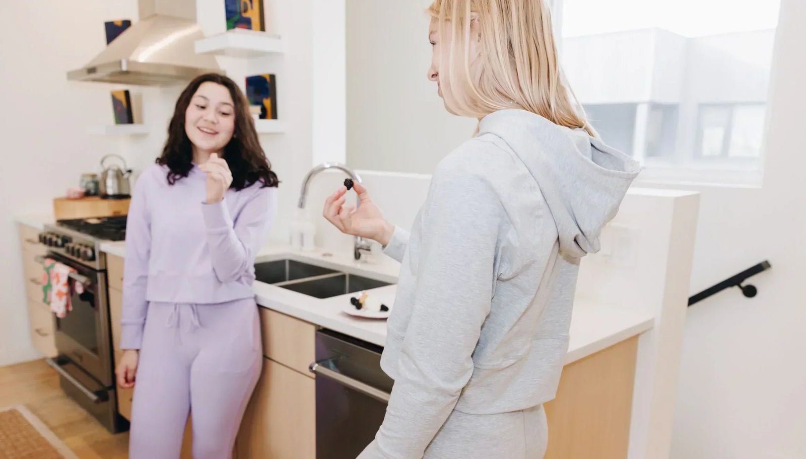 Two girls standing in a kitchen talking and eating, each one is wearing a matching lounge set. One set is huckleberry colored, the other set is grey colored.