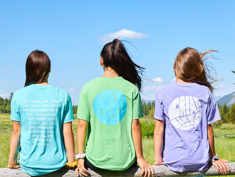 Three Girls sitting on a wooden fence facing away from the camera. Each one is in a different brightly colored t-shirt and it is a beautiful, blue sky summer day.