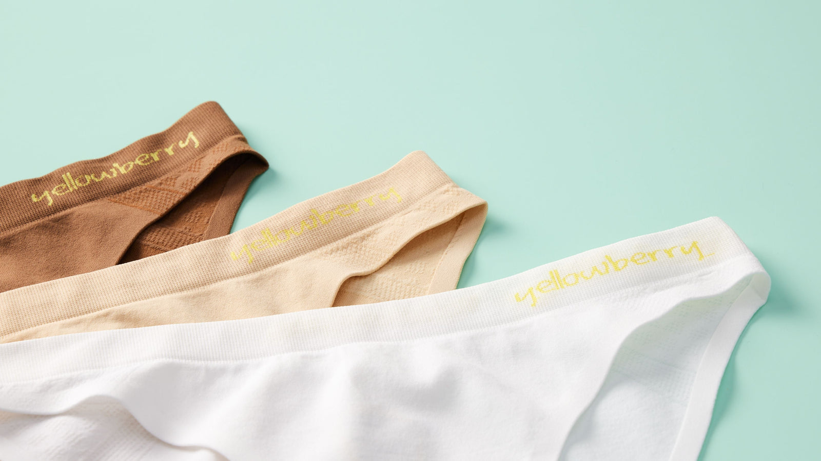 Three pair of Yellowberry Seamless Underwear, one mocha color, one doe color, one snowflake color. They are displayed on a soft green color background.