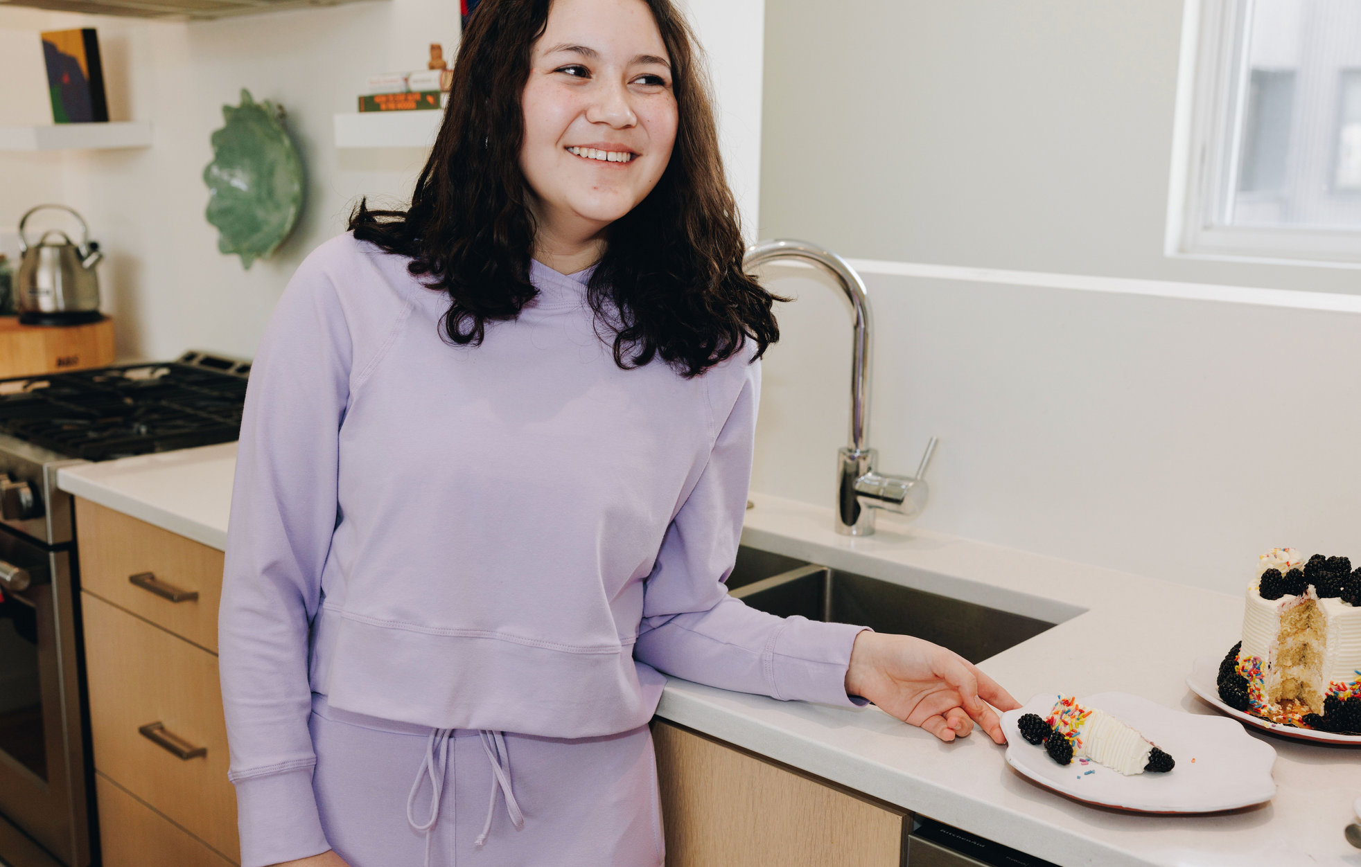 Girl laughing and standing at a counter top with a plate of cake. She is wearing a matching set of Yellowberry Downtown hoodie and Uptown Joggers in huckleberry.