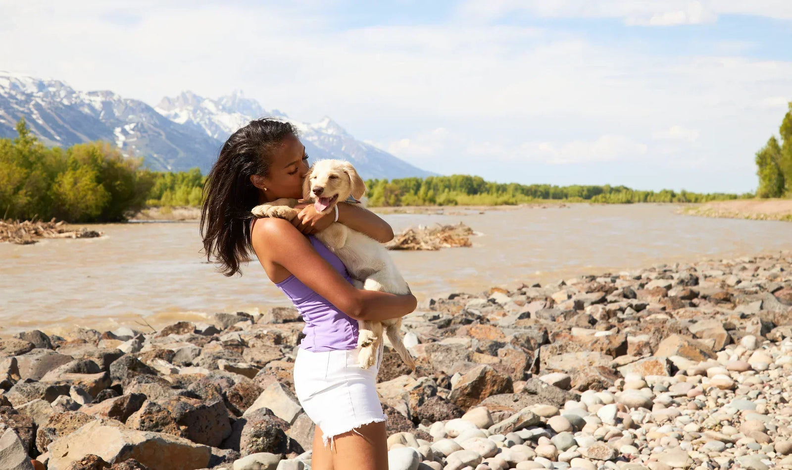 Girl standing next to a river and beautiful mountains in the distance holding an adorable puppy. She is wearing a Yellowberry Camisole while she kisses the puppy. It's a sunny, summer day.