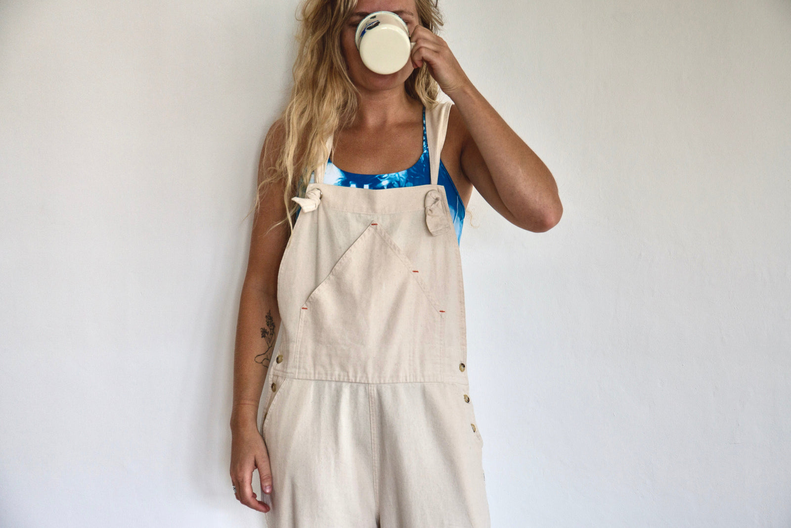 Girl wearing a blue yellowberry bra, peeking out from behind her adorable white overalls. She is sipping a drink.