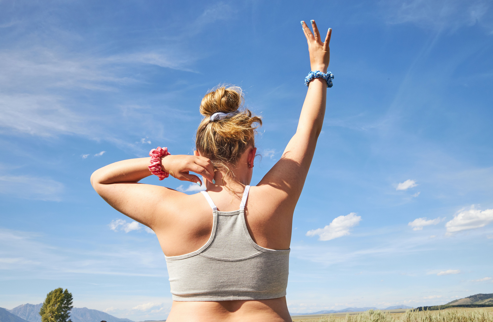 Girl standing against a blue sky. She is facing away from the camera and wearing a grey Yellowberry Bra. She has one hand in the air and colorful scrunchies on her wrist. It is a sunny day outside.