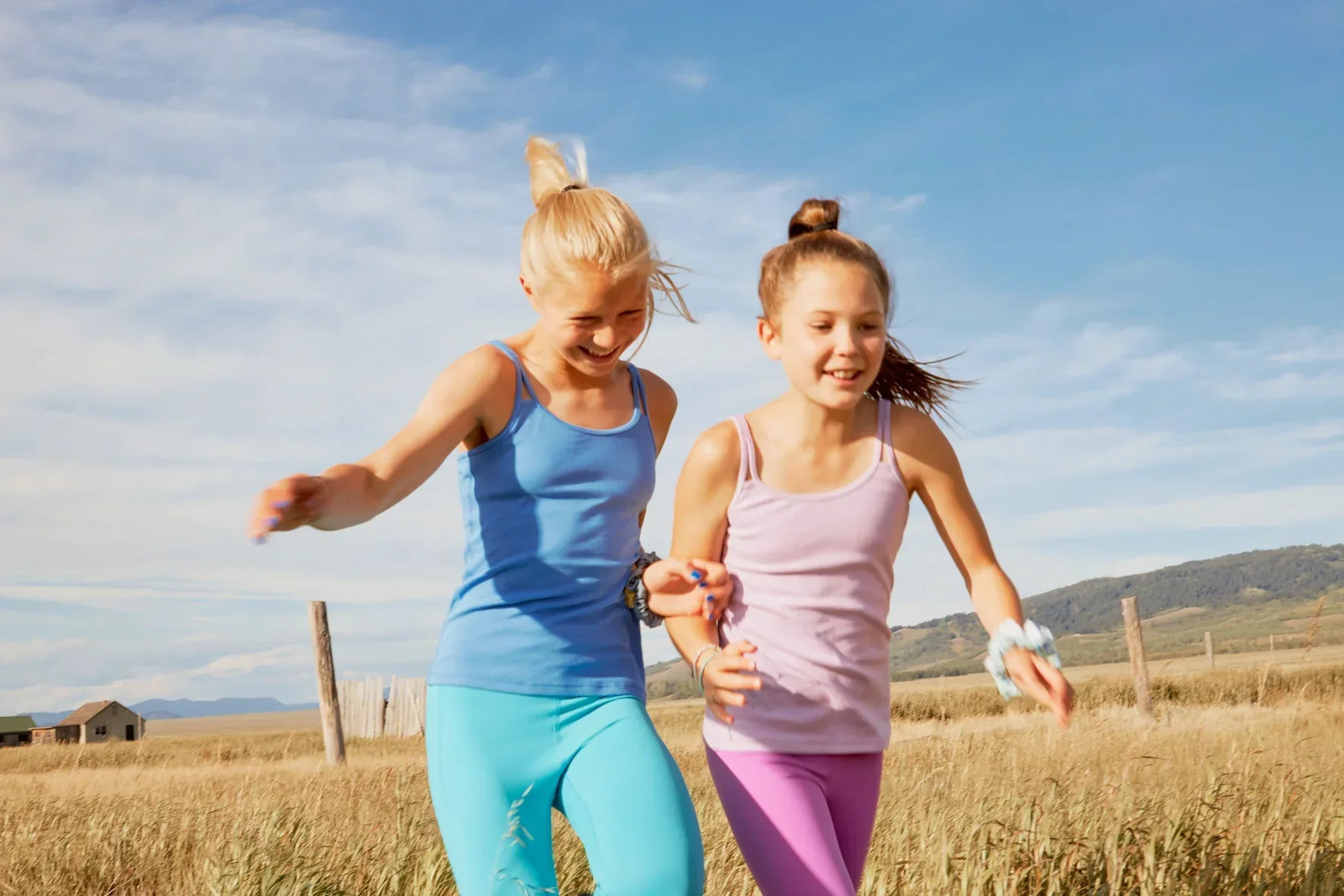 Two girls wearing Yellowberry tank tops and leggings laughing and running in the tall grass