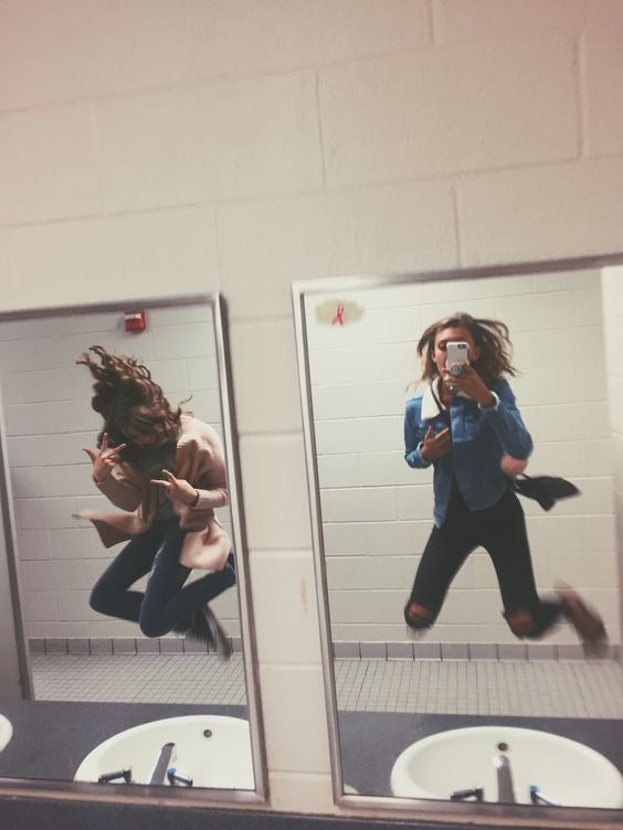 Two girls jumping up for a photo in front of a sink and mirror in the bacthroom