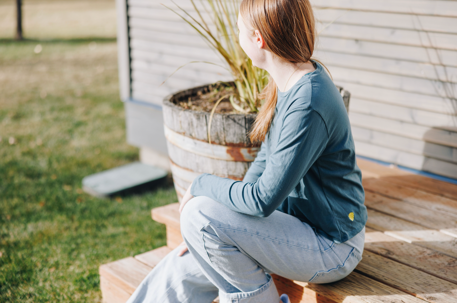 Girl sitting on a wooden deck with an elbow on her knee. she is wearing jeans and a turquoise cotton long-sleeve t-shirt by Yellowberry