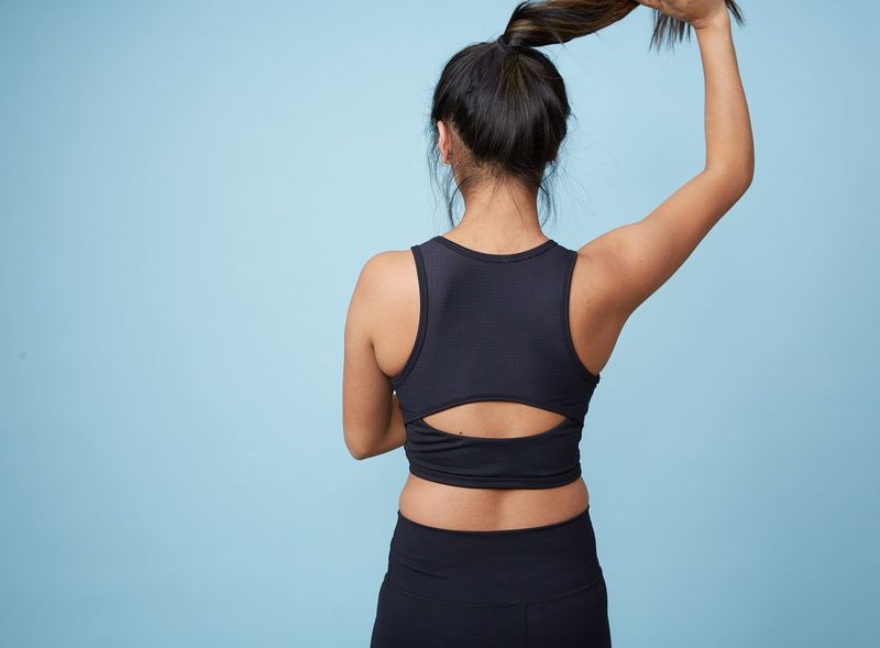 Girl standing against a blue backdrop, facing away from the camera. She is wearing a black crop top and matching black leggings