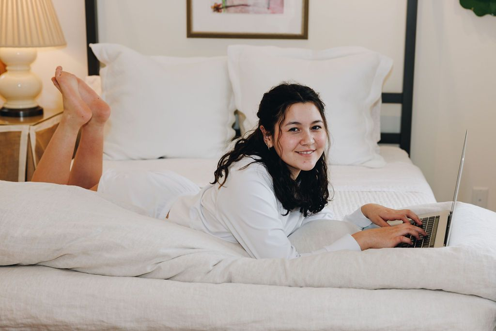 Girl lounging in bed typing on her computer. She is smiling at the camera and wearing a white cotton hoodie with matching white cotton shorts