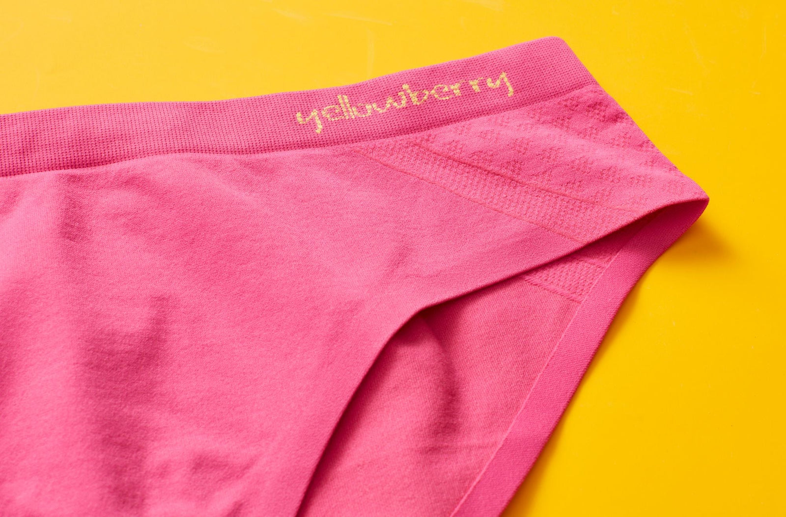 Closeup image of a pair of watermelon pink seamless underwear on a yellow background. The embroidered word "yellowberry" is written on underwear in yellow yarn 