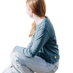 Girl wearing a blue green long sleeved cotton t-shirt sitting down with her elbow on her knee looking away from the camera