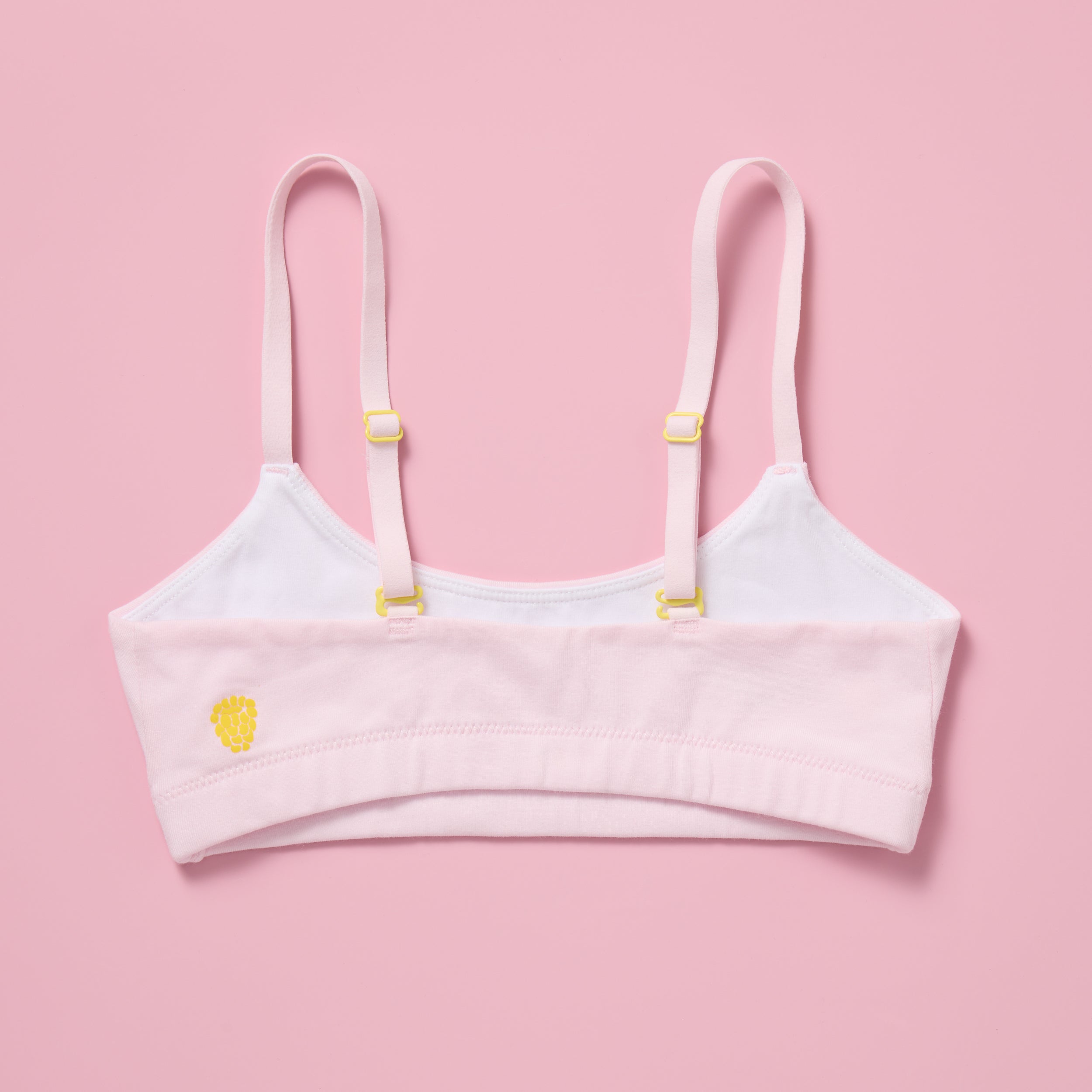 Yellowberry Pipit Training Bra for girls in Bubblegum (light pink) laid flat on a pink background, showing the back with adjustable convertible straps, yellow hardware, and the Yellowberry logo embroidered on the band
