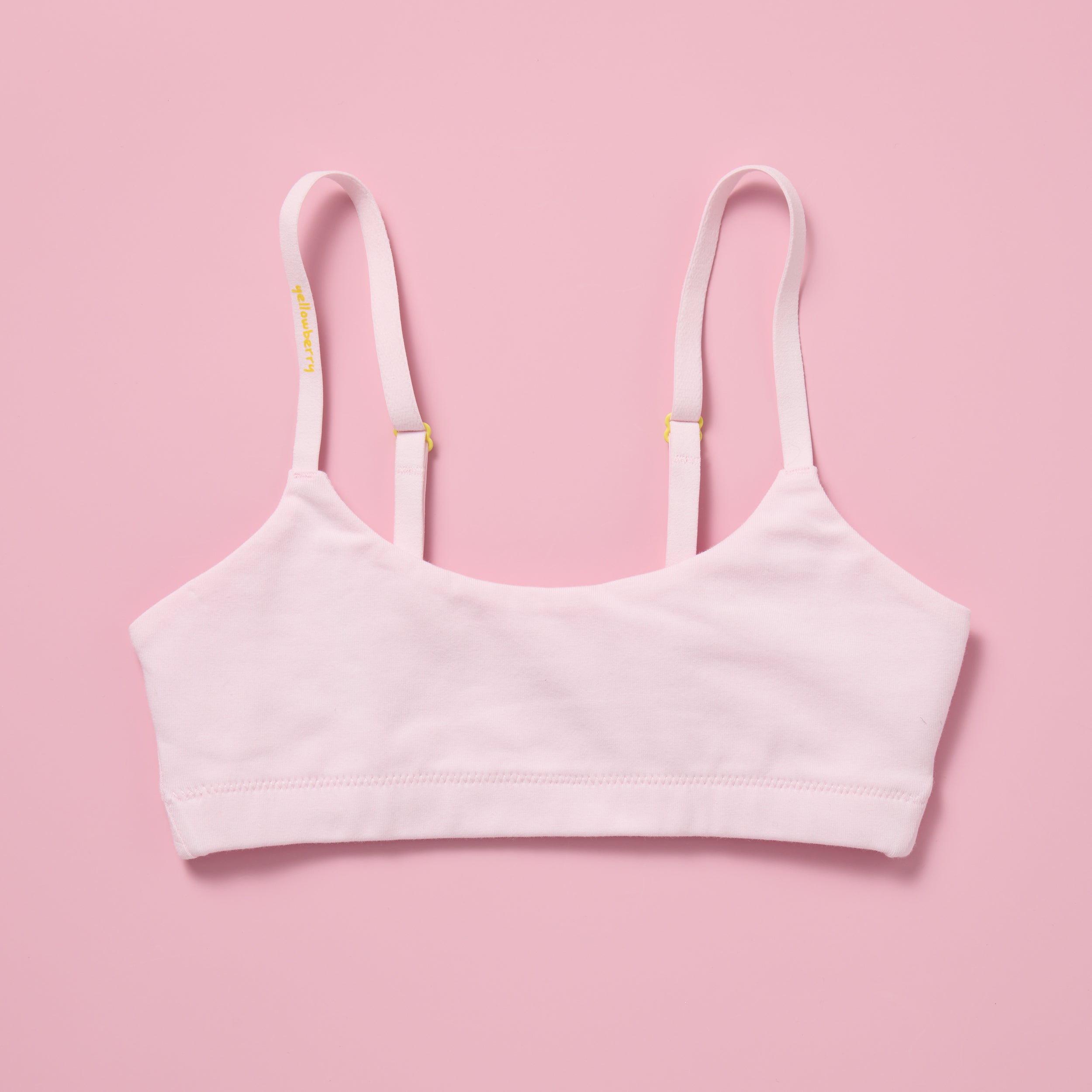 Yellowberry Pipit Training Bra and First Bra for girls in ultra soft Bubblegum (light pink)  fabric laid flat on a pink background, showing the front with adjustable convertible straps and yellow hardware
