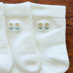 Three pairs of white knit socks arranged on a wooden surface, each featuring a small embroidered floral motif near the cuff in pale yellow and green.