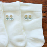 Three pairs of white knit socks arranged on a wooden surface, each featuring a small embroidered floral motif near the cuff in pale yellow and green.