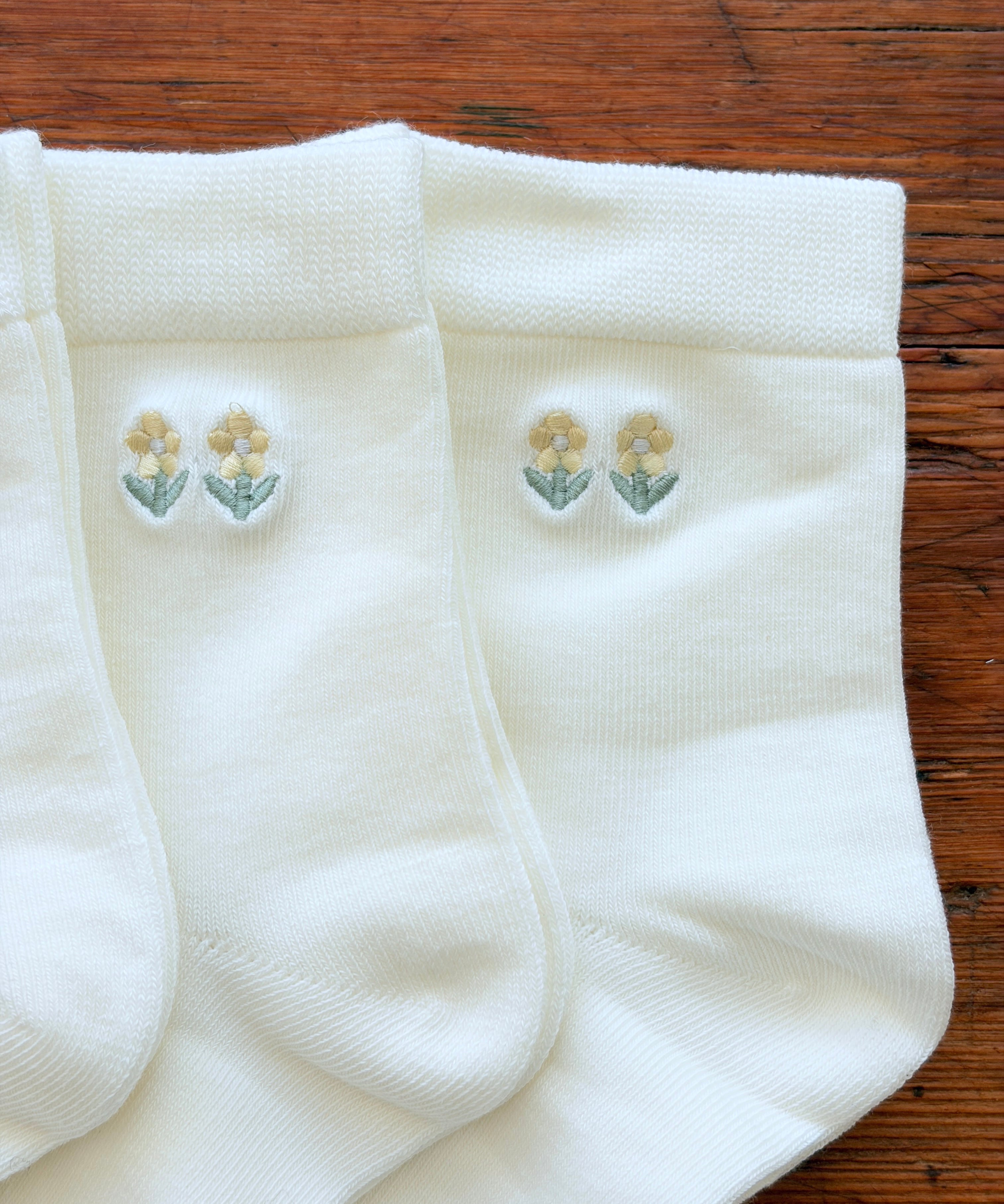 Three pairs of white knit socks arranged on a wooden surface, each featuring a small embroidered floral motif near the cuff in pale yellow and green.