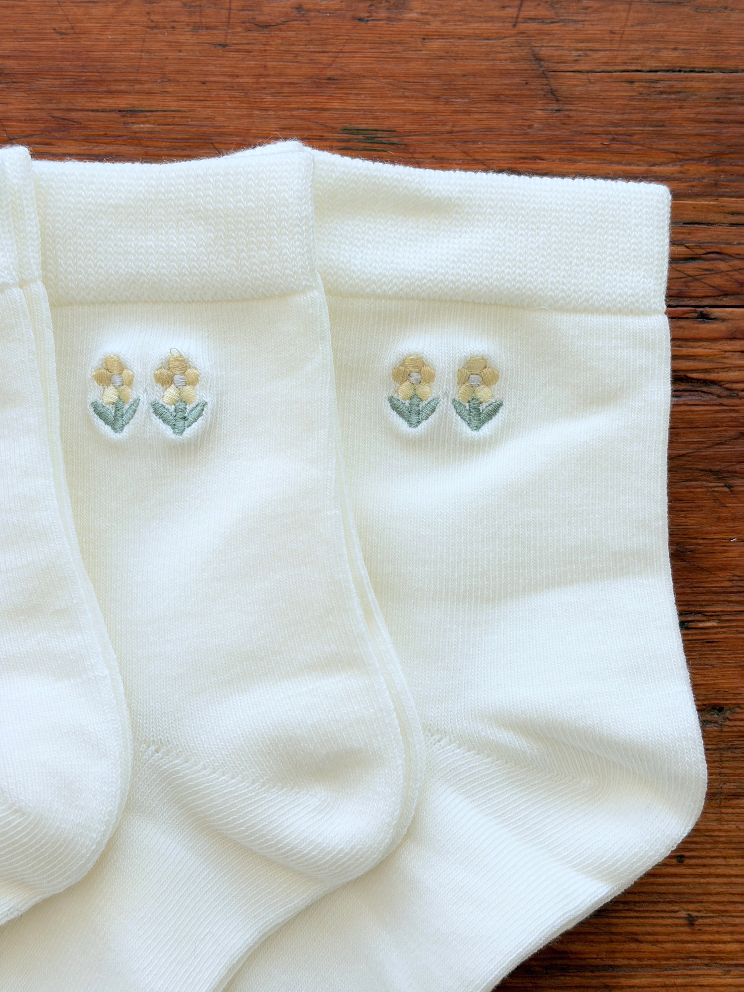 Three pairs of white knit socks arranged on a wooden surface, each featuring a small embroidered floral motif near the cuff in pale yellow and green.
