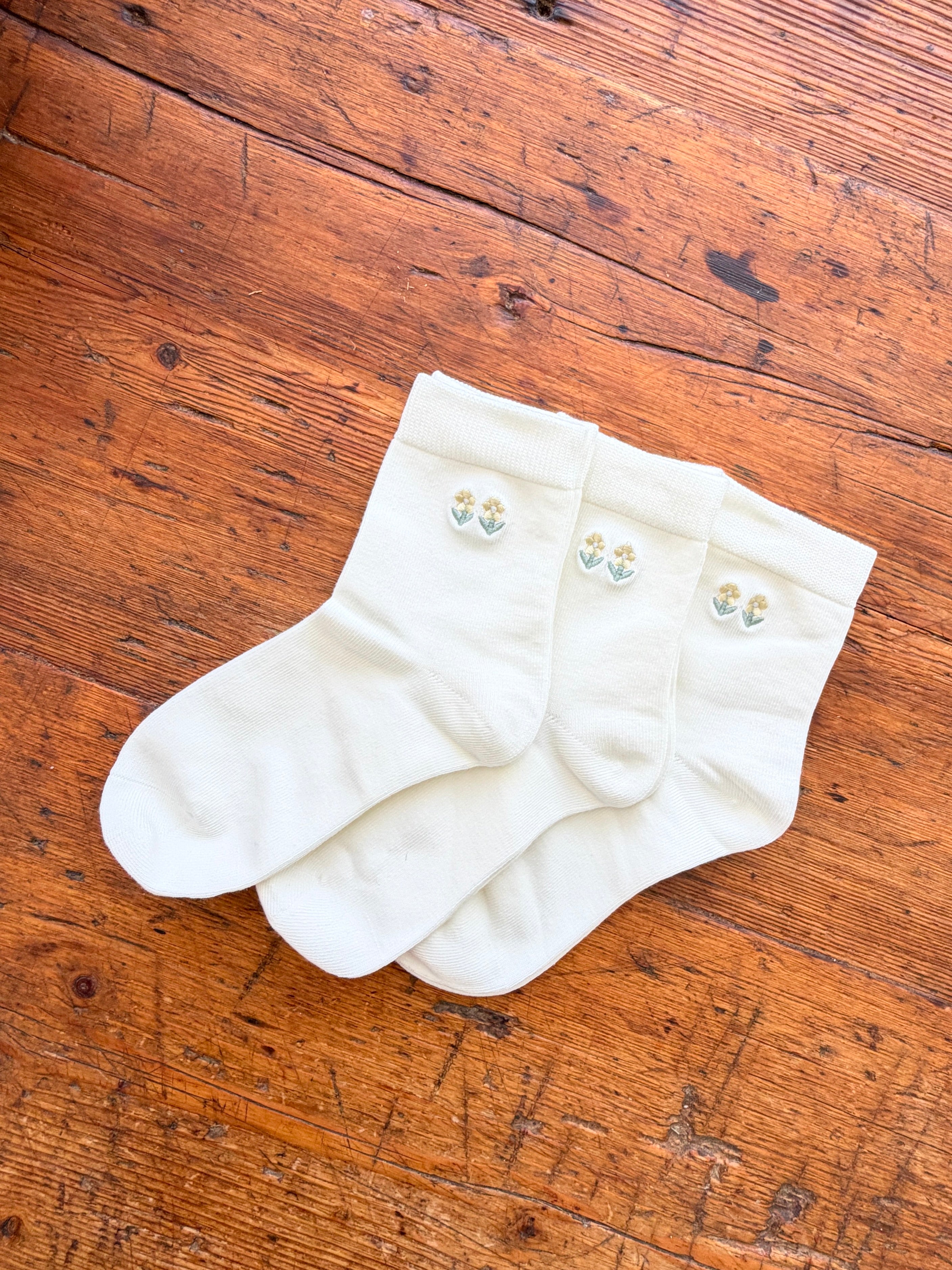 Three off-white crew socks with small embroidered tulip motifs in light blue and yellow, arranged on a rustic wooden surface.