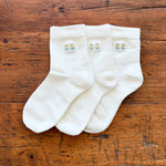 Three white crew socks with small embroidered floral motifs on the cuffs, arranged on a rustic wooden surface.