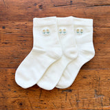 Three white crew socks with small embroidered floral motifs on the cuffs, arranged on a rustic wooden surface.
