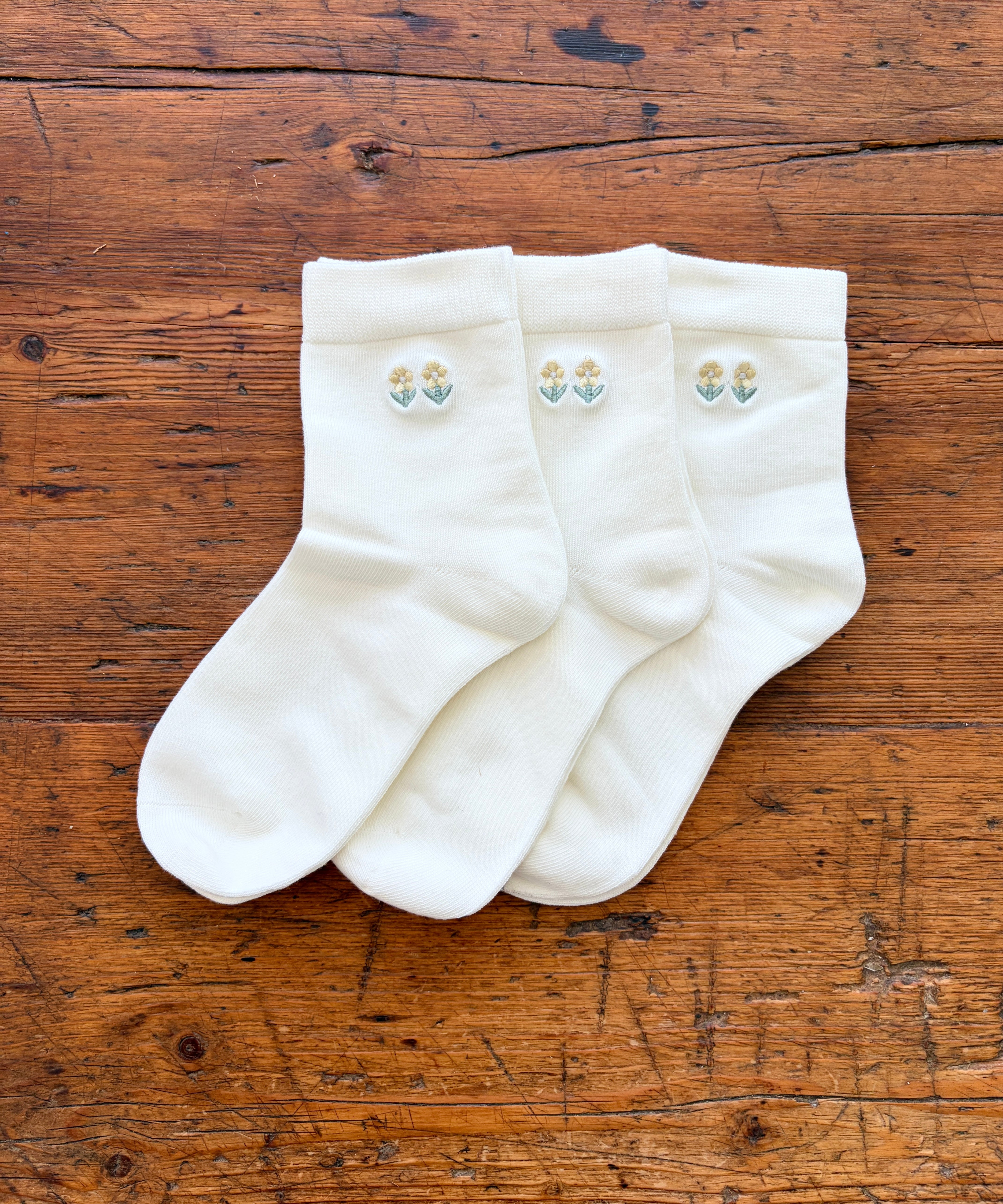 Three white crew socks with small embroidered floral motifs on the cuffs, arranged on a rustic wooden surface.