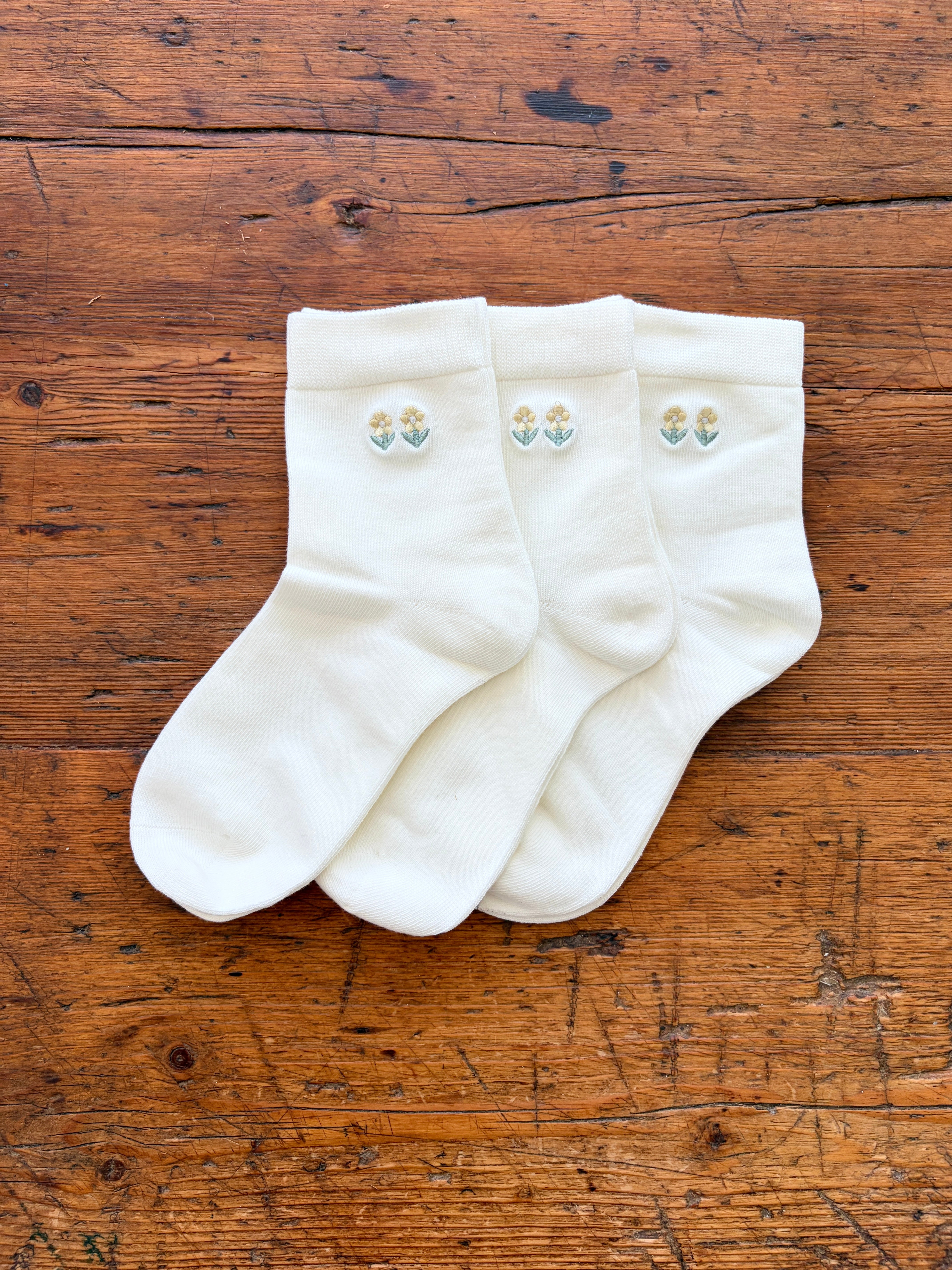 Three white crew socks with small embroidered floral motifs on the cuffs, arranged on a rustic wooden surface.