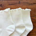 Close up of three pairs of white ribbed ankle socks with ruffled edges, arranged on a rustic wooden surface.