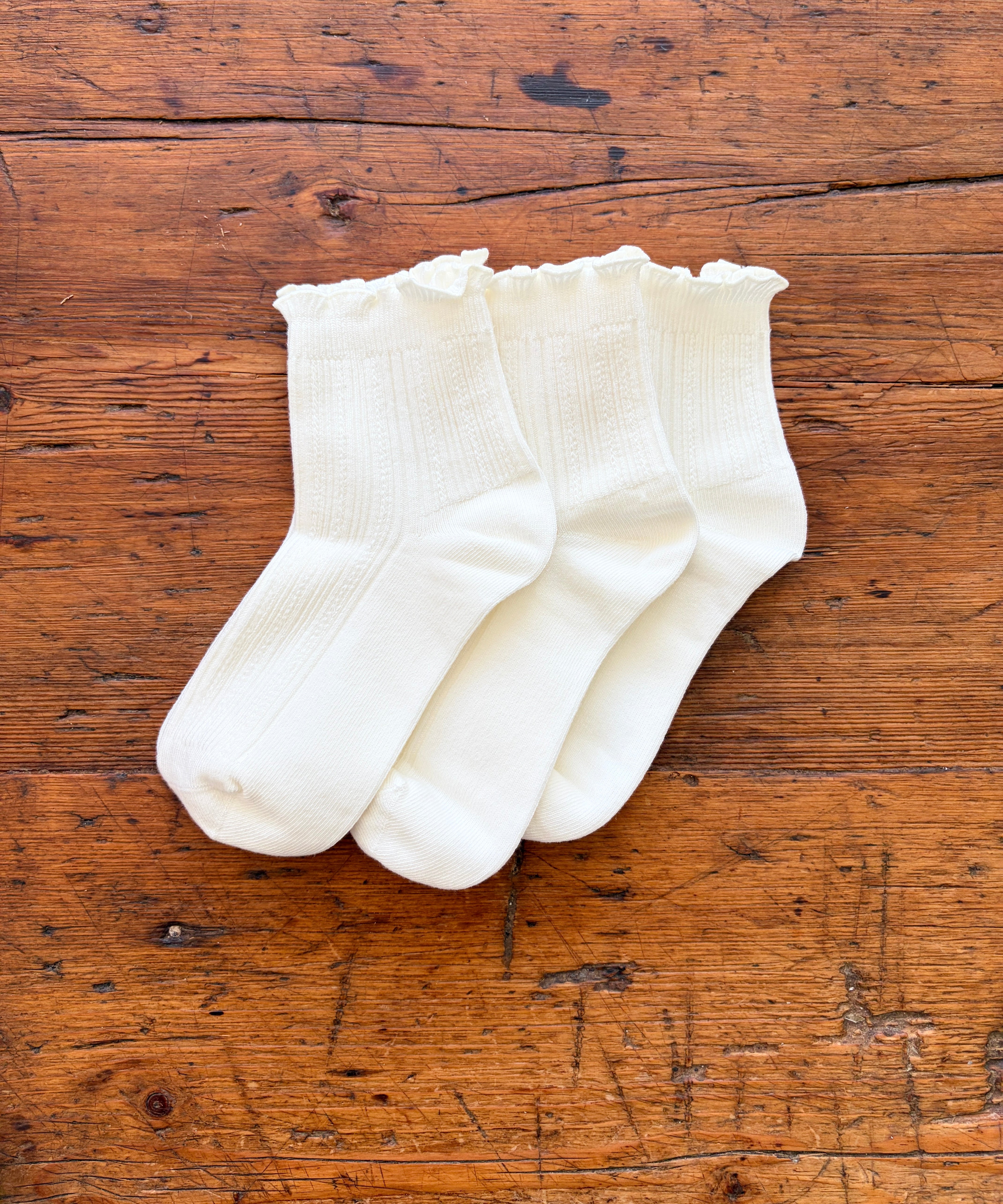 Three pairs of white ankle socks with ruffled edges, arranged on a rustic wooden surface.