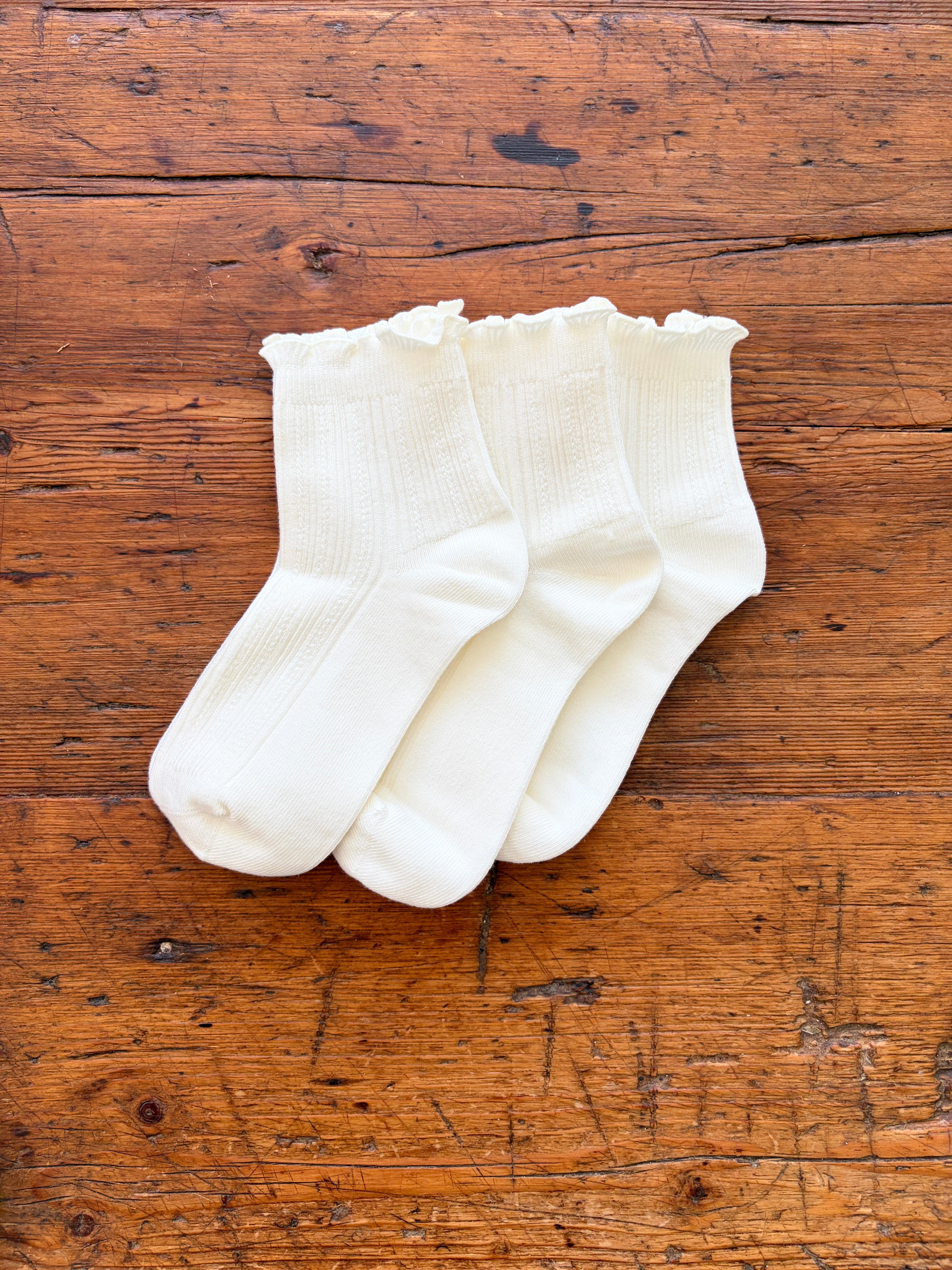 Three pairs of white ankle socks with ruffled edges, arranged on a rustic wooden surface.