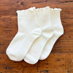 Three pairs of white ribbed ankle socks with ruffled edges, arranged on a rustic wooden surface.