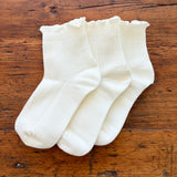 Three pairs of white ribbed ankle socks with ruffled edges, arranged on a rustic wooden surface.