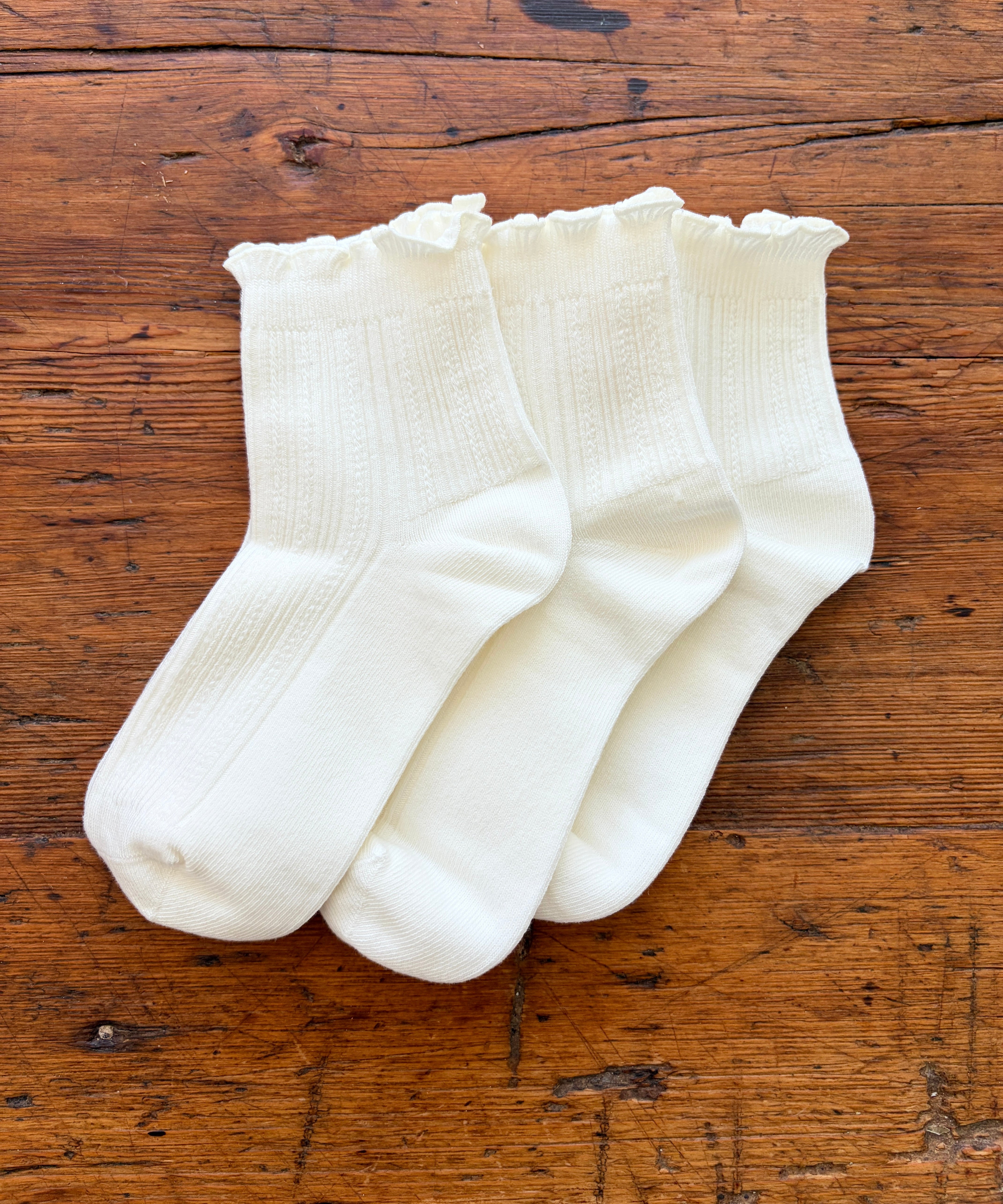 Three pairs of white ribbed ankle socks with ruffled edges, arranged on a rustic wooden surface.