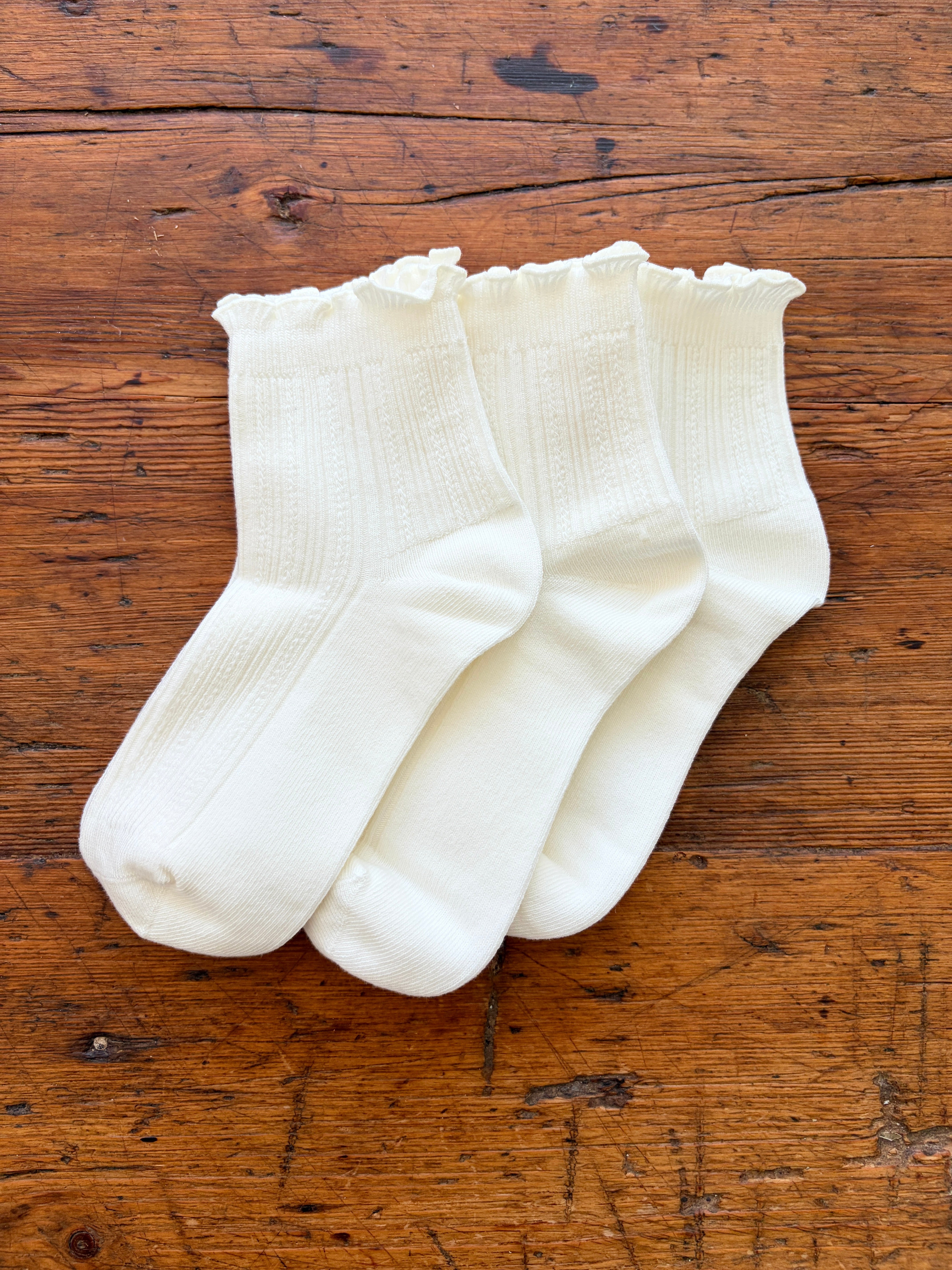 Three pairs of white ribbed ankle socks with ruffled edges, arranged on a rustic wooden surface.