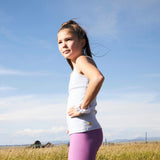 Girl is standing from the side, looking up. She is wearing a light blue, shelf cotton camisole with a shelf bra as well as purple leggings and a scrunchie on her wrist. Her hair is in a ponytail and there is a blue sky behind her.