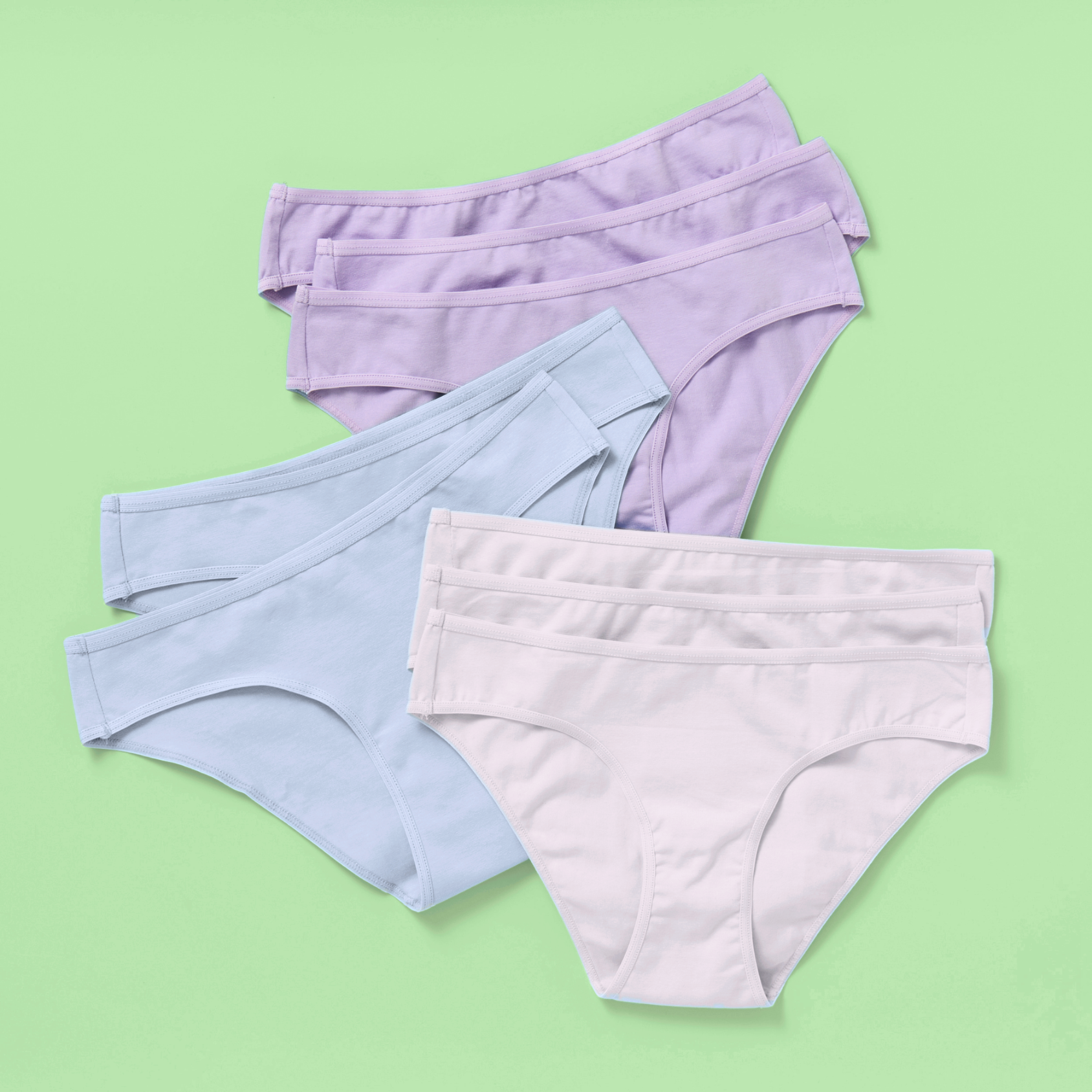 Eight pairs of women's high-waisted briefs in pastel shades of lavender, light blue, and off-white, arranged in a stacked fan layout on a solid light green background.