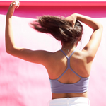 A woman viewed from behind wearing a lavender sports bra with a crisscross open-back design, set against a bright pink background.